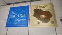 Two vintage metal bar signs, left Bacardi blue sign, right Hennessy Cognac cream colored sign on tile floor.
