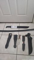 Photo of 8 knives and a machete with their sheaths, laid out on tiled floor.
