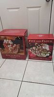 Photo showing two original red boxes side by side on the floor, each box features an image of the corresponding Fitz and Floyd holiday collectible inside.