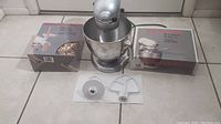 Full view of the KitchenAid Artisan mixer with boxed pasta maker and pouring shield and three mixer attachments laid out in front.