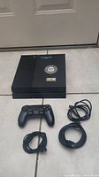 Photo of black PS4 console with Weedmaps and MFXX stickers along with one controller and cords on tiled floor.