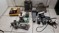 Overall view of the collection of vintage cameras and projectors laid on white tiled floor, showing multiple models including box cameras, flash units, and projectors.