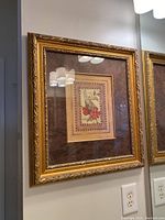 Frontal angled photo of one framed botanical print showing ornate gold frame with glass and reflections. Brown patterned mat and cream illustrated flower artwork inside.