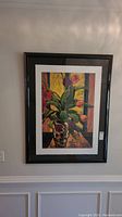 Framed signed art print of tulips in a vase hung on a wall. Colorful painting with yellows, greens, pinks, and purples, in a dark wood or black frame with white matte border.