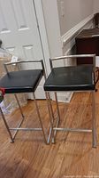 Two bar stools with black faux leather square seats and chrome metal bases, positioned on wood flooring near a white door.