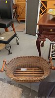 Oval wicker basket with visible wooden handles, placed on floor among other furniture.