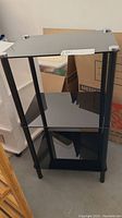 Front angled view of black metal shelf unit with three shelves in a room with boxes and storage items.