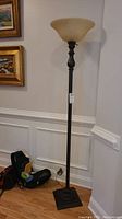 Full view of bronze metal floor lamp standing on hardwood floor against white wall with molding. Square base and tall turned post design, bronze finish.