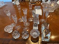 Wide view showing sixteen varied crystal and glass candle holders and decorative pieces on wooden table.