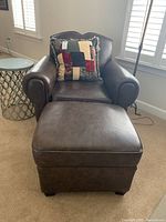 Full view of the brown leather club chair with rolled arms, the matching ottoman, and the patchwork throw pillow on the chair.