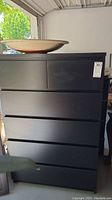 Front view of black chest of drawers showing six drawers and some surface scratches and dust.
