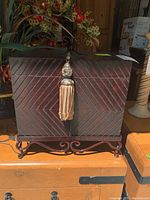 Front view of wooden box showing chevron design, decorative tassel, and metal stand.