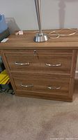 Front view of two-drawer wood file cabinet with lock and key in top drawer, showing metal handles and wood finish. Lamp base on top surface visible.