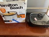 Hamilton Beach slow cooker in box next to Bella electric skillet with glass lid, all placed on wooden table.