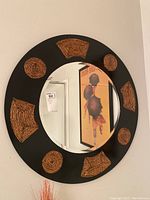 Full front view of round mirror showing black frame and multiple woven accents in circular and polygon shapes around mirror glass.