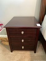 One red mahogany nightstand showing front view of three drawers with round silver knobs, wood surface glossy with scratches.