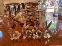 Four glass rabbit figurines with colored ears and tails placed in front of a detailed wooden model of the Mayflower ship with sails and rigging on a wooden table.