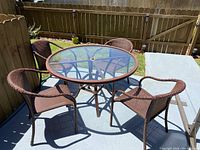 Full view of the round glass table with metal base and four brown woven armchairs arranged outdoors.
