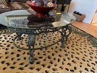 View of the oval coffee table showing the full glass surface, beveled edges, and metal base with intricate scroll detail and claw feet.