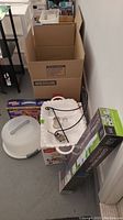 View of boxed kitchen and serving items including white plastic cake cover and various boxed tray and pitcher sets, placed on floor near cardboard packing boxes.