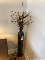 Full view of tall floor vase with faux wooden sticks arranged inside, showing overall shape and height