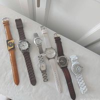 7 wristwatches aligned side by side on a white cushioned surface with fullbands shown, illustrating variety in color and style of the lot