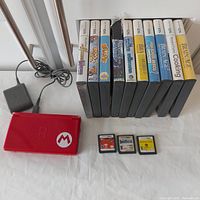 Photo showing red Nintendo DS Lite console closed with Mario logo, charger, and 13 DS games (10 in cases) lined up vertically