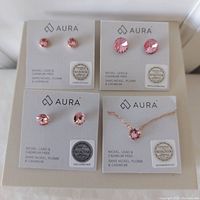Four pieces of Swarovski crystal jewelry including three pairs of rose gold plated pink crystal stud earrings and one matching rose gold plated necklace with pink crystal pendant, shown mounted on Aura brand cards with Swarovski logos and authenticity seals.