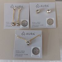 Photo showing three Aura branded jewelry pieces with Swarovski crystal authenticity stickers: two sets of earrings and one necklace with clear crystals, arranged on white display cards.