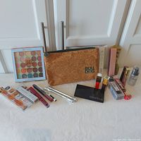 Full view of the entire cosmetics lot displayed showing the cork look Bull Dog bag, eyeshadow palette, lipsticks, mascaras, and various boxed items.