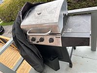 Full view of Chef Master propane BBQ grill with black worn cover partially draped over it. Grill has three knobs and a side shelf.