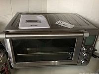 Front view of Breville stainless steel toaster oven showing glass door, control panel, and power cord. User manual placed on top.