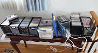 Photo showing six black wire bins with CDs and some cassettes, two blue lidded plastic bins with DVDs, and two white extension cords arranged on a wooden table.