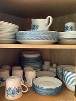 Shelf of Corelle plates, bowls, and mugs with blue stripe pattern and blue floral details shown together.