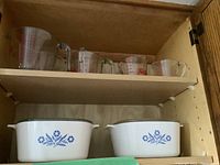 Photo shows two white Corning Ware covered casseroles with blue cornflower design on bottom shelf, and five clear Fire King glass measuring cups with red and blue markings arranged on upper shelf inside wooden cabinet.