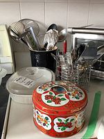 Photo showing black utensil holder filled with various kitchen utensils, white Corning Ware oval bakeware with lid, decorative floral tin container, and a small metal utensil holder containing flatware.