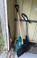 Three garden tools leaning against a wooden shed wall: green plastic leaf rake, black plastic garden rake, and shovel with wooden handle and D-grip