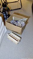 Box on the floor with various sealed plastic bags presumably containing cabinet hardware pieces beside a wicker basket.