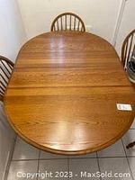 Full view of the oak kitchen table showing the oval wooden top and a portion of chairs around it.
