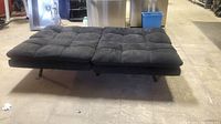 Photo of the futon in fully flat position as a bed, showing tufted black fabric upholstery and black metal legs.
