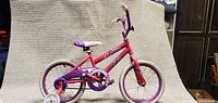 Full side view of the pink and purple kids bike with training wheels, showing frame, wheels, chain protector, and seat.
