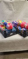 Three black crates filled with multiple colorful plastic balls for kids arranged on a beige textured fabric surface.