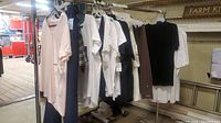View of various new clothing items hanging on metal clothes rack, including white tees, black top, brown pants, blue pants and light pink shirt.