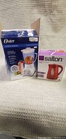Photo showing the Oster 12-Speed Blender box and Salton red kettle box side by side on a fabric surface.