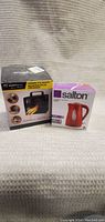 Photo of the boxed red Salton kettle and ceramic heater placed side by side on fabric background.