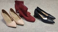 Three pairs of women's shoes on a beige fabric background. Beige closed-toe heels, red suede ankle boots, black crocodile texture mid-heel pumps.