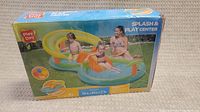 Box front showing Play Day Splash & Play Center with image of children and adult using the inflatable pool featuring a slide and splash arch.