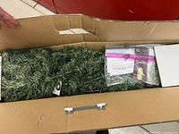 View into the open box showing green plastic pine tree sections and plastic bag with instructions and hardware.