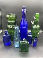 Lot displayed with 9 glass bottles in cobalt blue and green, various shapes and sizes, including apothecary and beer bottle styles.