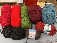 Assorted skeins of yarn in various colors such as red, green, maroon, blue, black, and brown on a gray table.